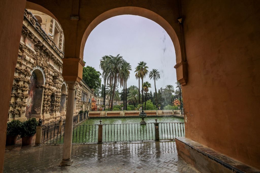 Looking into the gardens at the Real Alcazar with palm trees and a pool behind a yellow arch. 