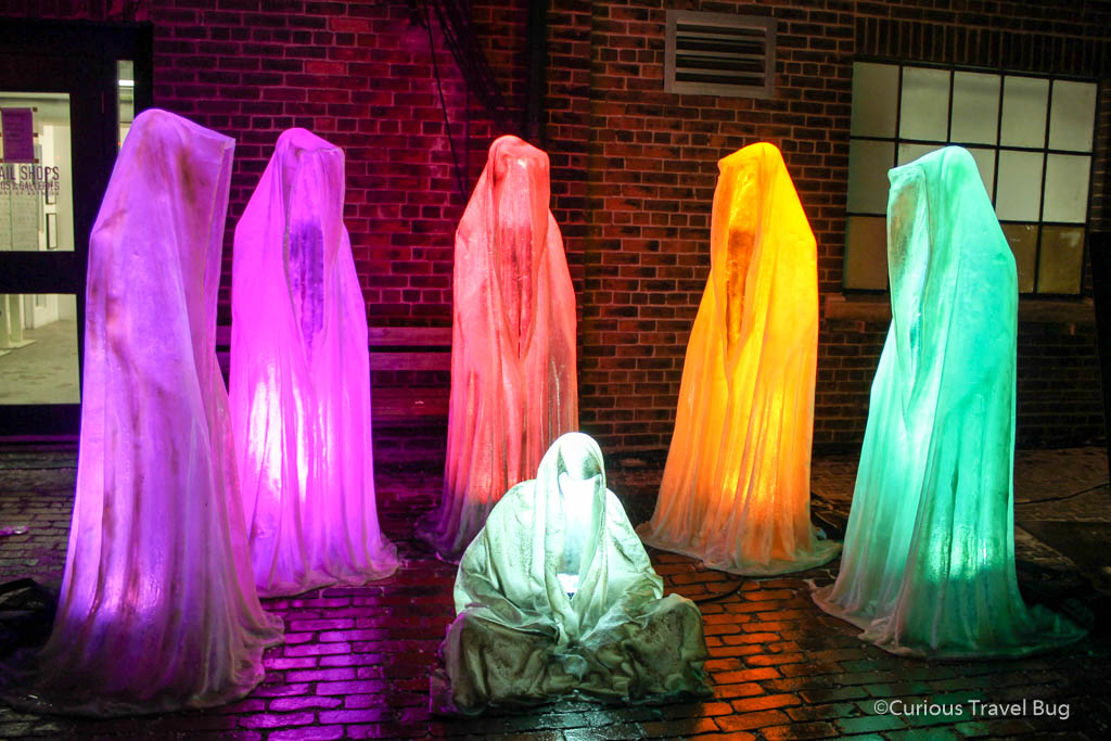 A light installation of five colourful shrouded figured standing around a figure sitting cross legged in Toronto's Distillery District during the Light Festival. It's one of the best things to do in Toronto during winter