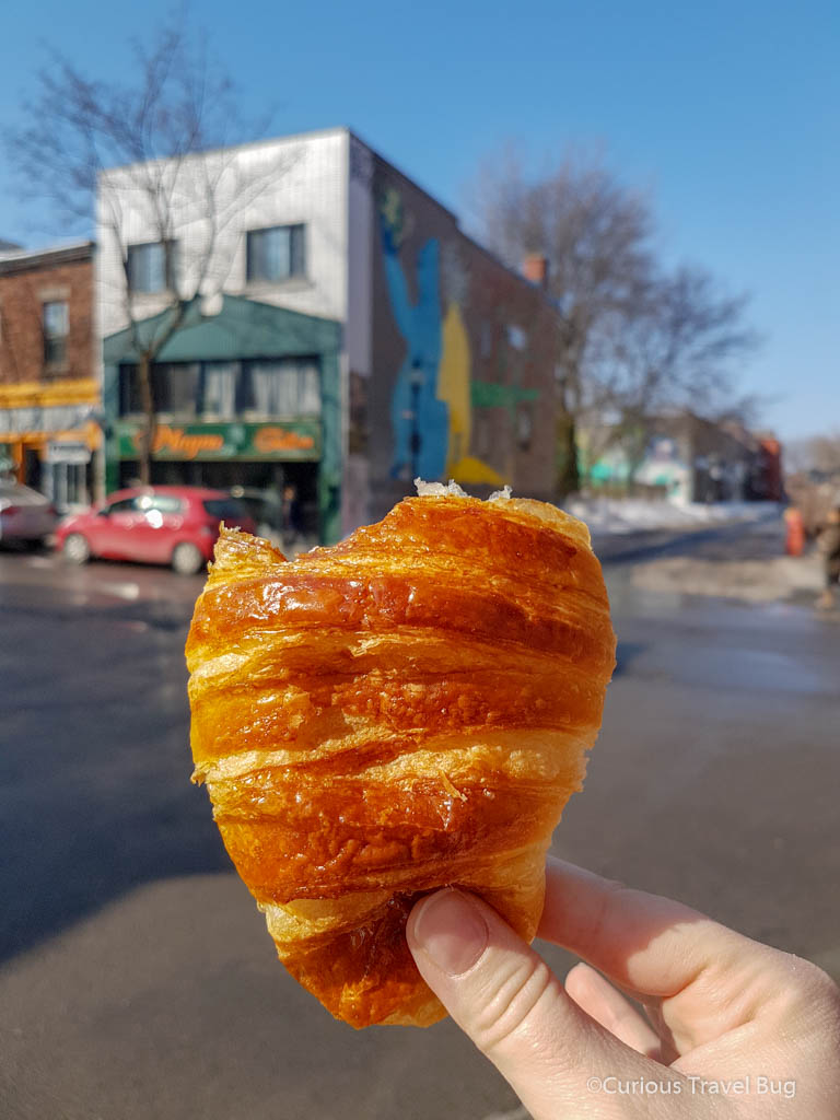 A croissant from Patisserie Kouign Amman in Montreal with the city behind it. This is an amazing French Bakery experience in Montreal that should not be missed. The croissants here are amazing and butter and flaky.