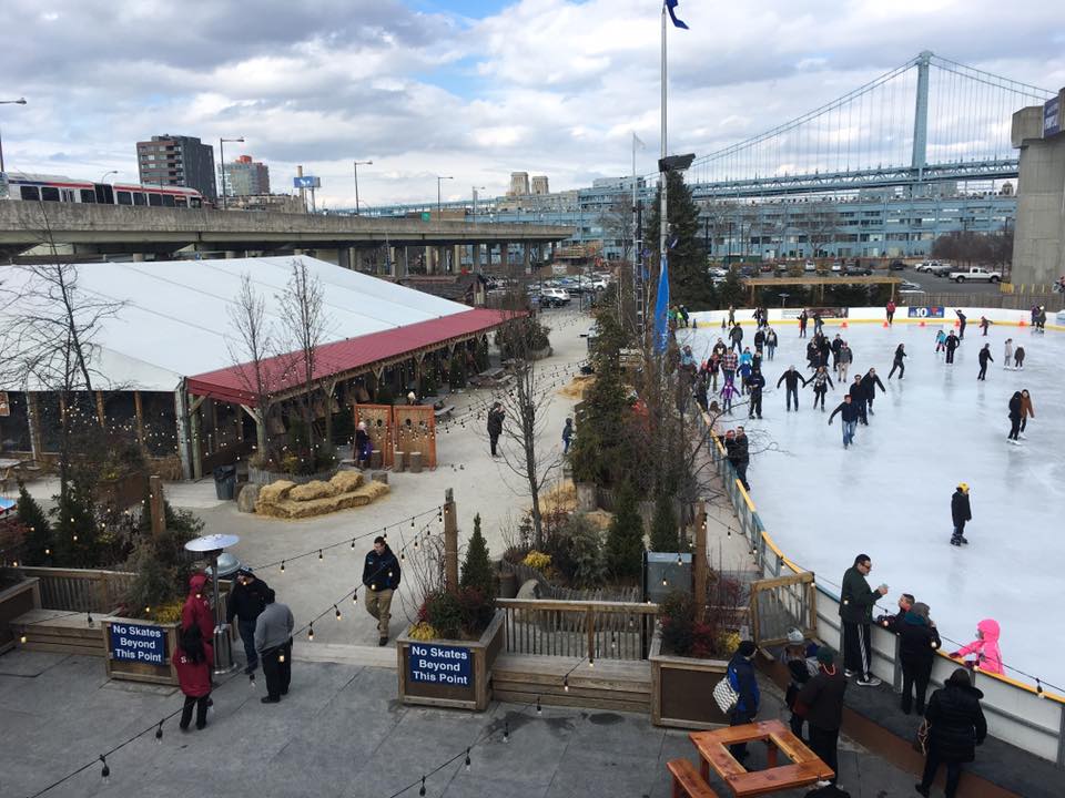 View of Philadelphia's Riverrink skating and winterfest with the bridge in the background. Blue Cross Riverrink is a great place to skate and perfect for any ice skating bucket list.