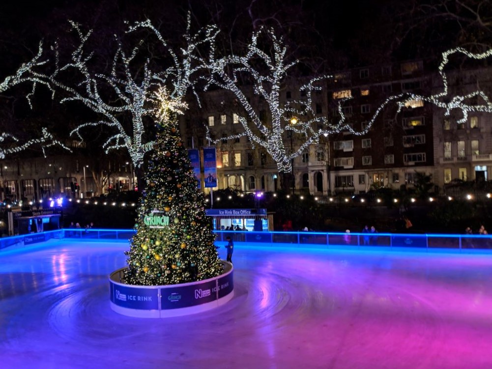 Skating rink in front of the London Natural History Museum with the ice lit up by colored lights and a Christmas Tree in the middle.
