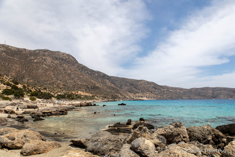 Clear water and a rocky beach with mountains in the background at Kedrodasos Beach in Crete. This beach is near to Elafonissi and makes for a great addition to any Western Crete itinerary