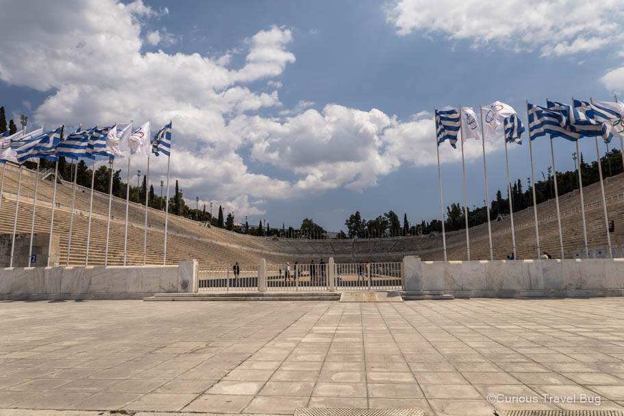 The Panathenaic Stadium where the first modern Olympic games were held with Olympic and Greek flags flying in front of it on a sunny day