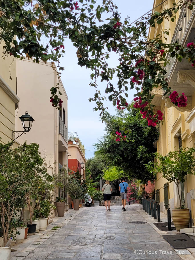 A couple walks in the streets of the Plaka near the Acropolis with colorful bougainvillea flowers overhead. This neighbourhood is perfect to walk through early morning while visiting Athens for four days.