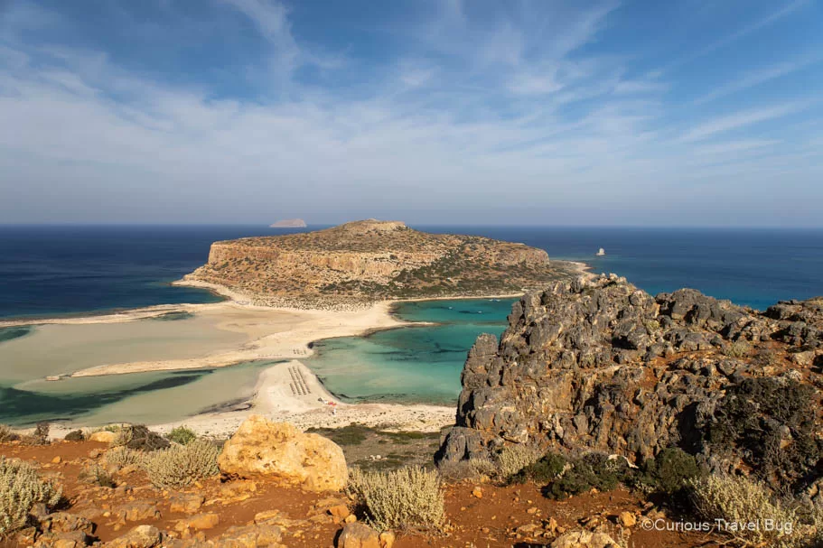 View of Balos Beach in Western Crete with clear blue skies and teal water, red soil and black rocks make up the forefront