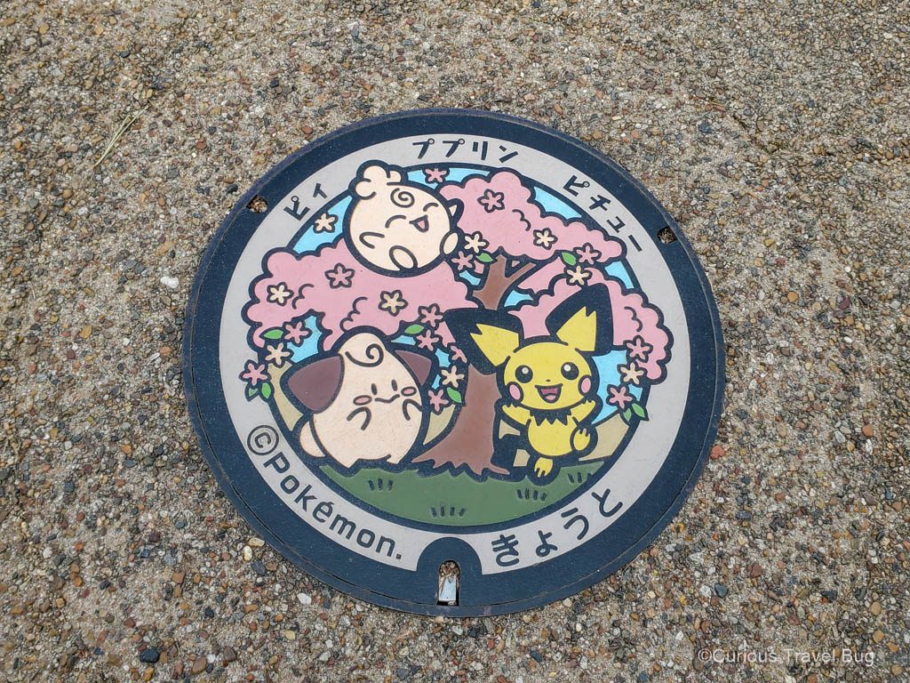 Manhole cover in Kyoto known as a pokelid because it is a colourful cover with the Pokemon igglybuff, pichu, and cleffa on it in front of a blooming cherry tree.